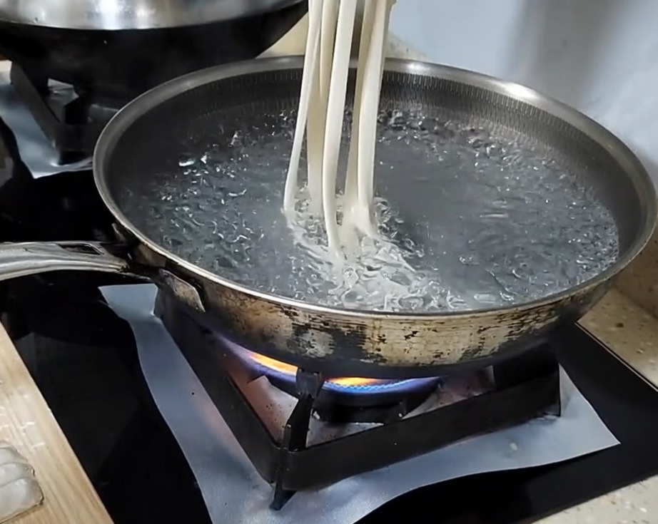 Hand stretching a dough strip into a long noodle before dropping it into a pot of boiling water.