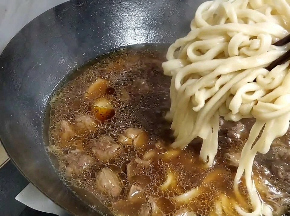 Thick noodles being lifted with chopsticks from a pot of water and placed into a wok with soup base.