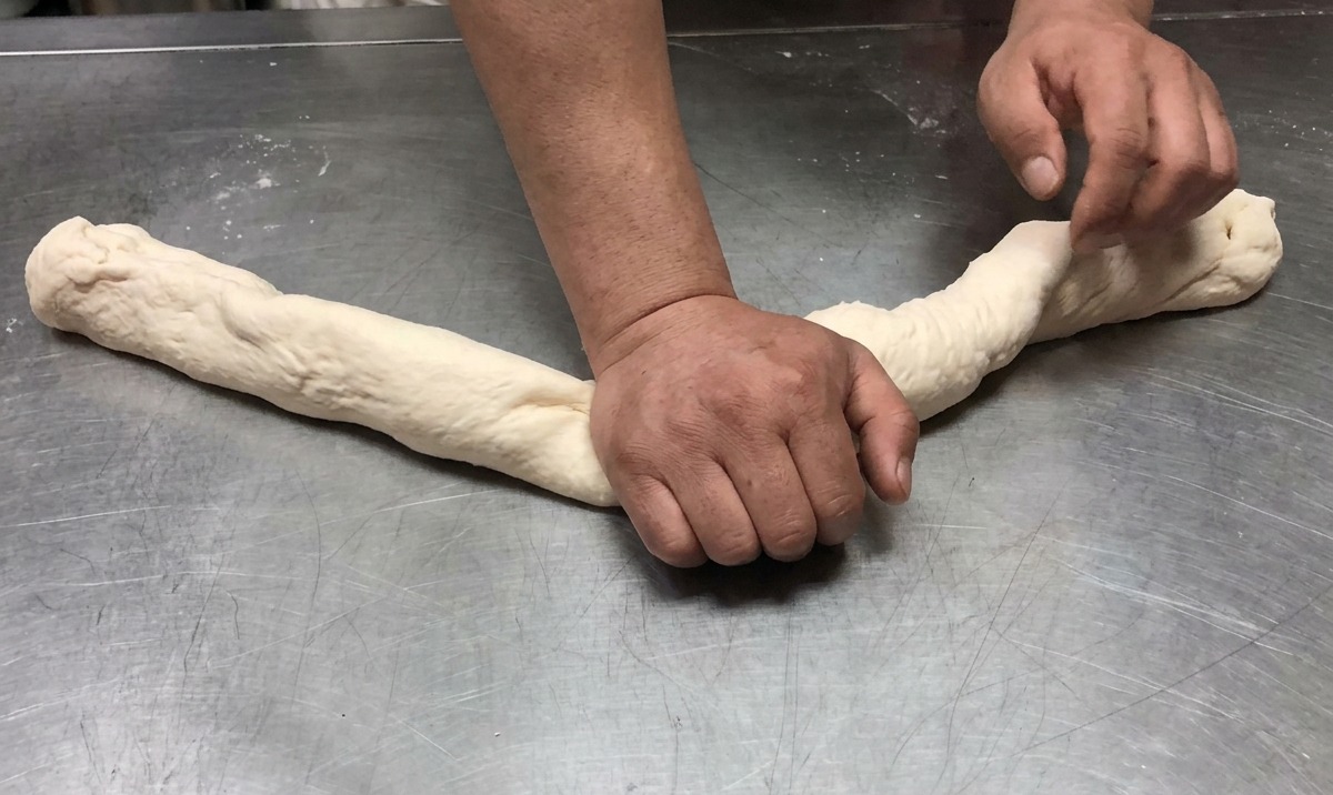 Dough being stretched and kneaded into a long rope on a stainless steel work surface.