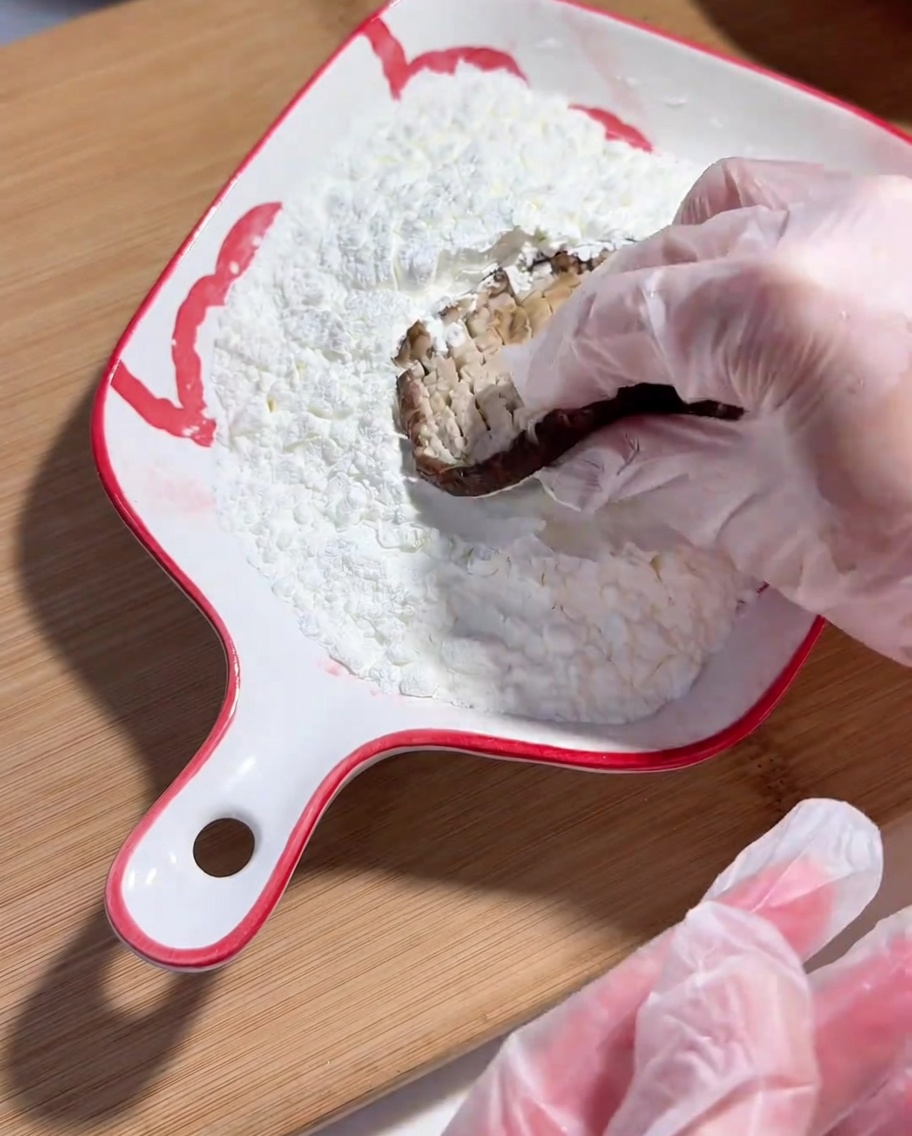 A hand wearing a plastic glove dipping the scored side of a shiitake mushroom into a dish of cornstarch.