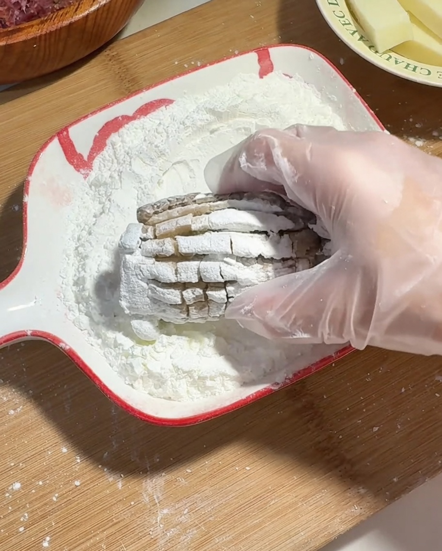 A hand wearing a plastic glove coating a beef and cheese stuffed shiitake mushroom in cornstarch.