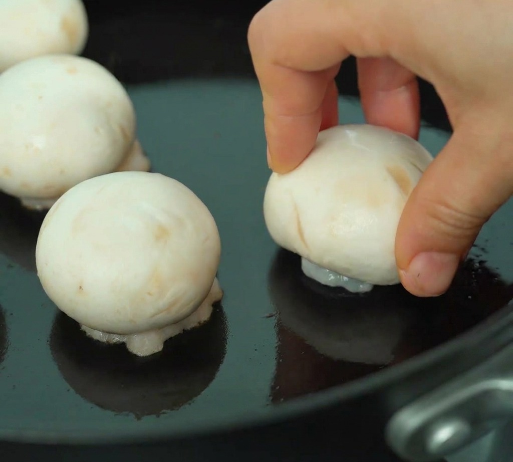 Shrimp-filled mushroom caps searing in a pan with the golden-brown shrimp side facing down.