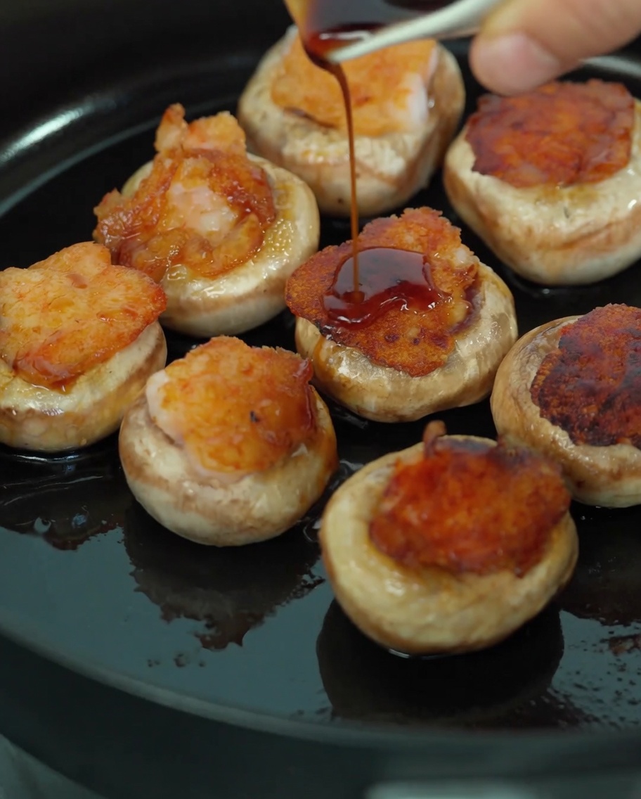 Pouring a dark soy sauce and mushroom juice mixture over shrimp-stuffed mushrooms in a pan.