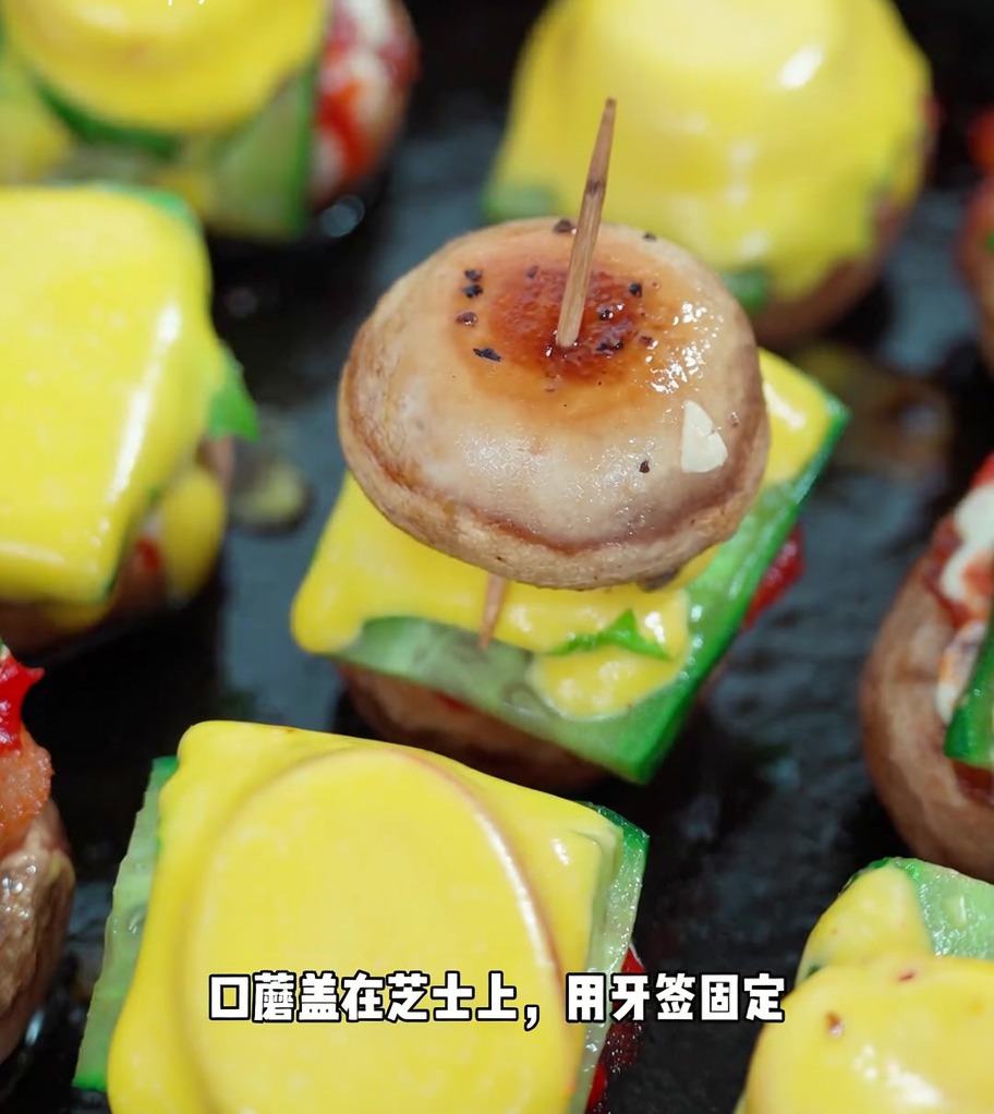 A finished mini mushroom burger secured with a toothpick, showing the stacked layers of mushroom, cucumber, lettuce, tomato, cheese, and a second mushroom cap.