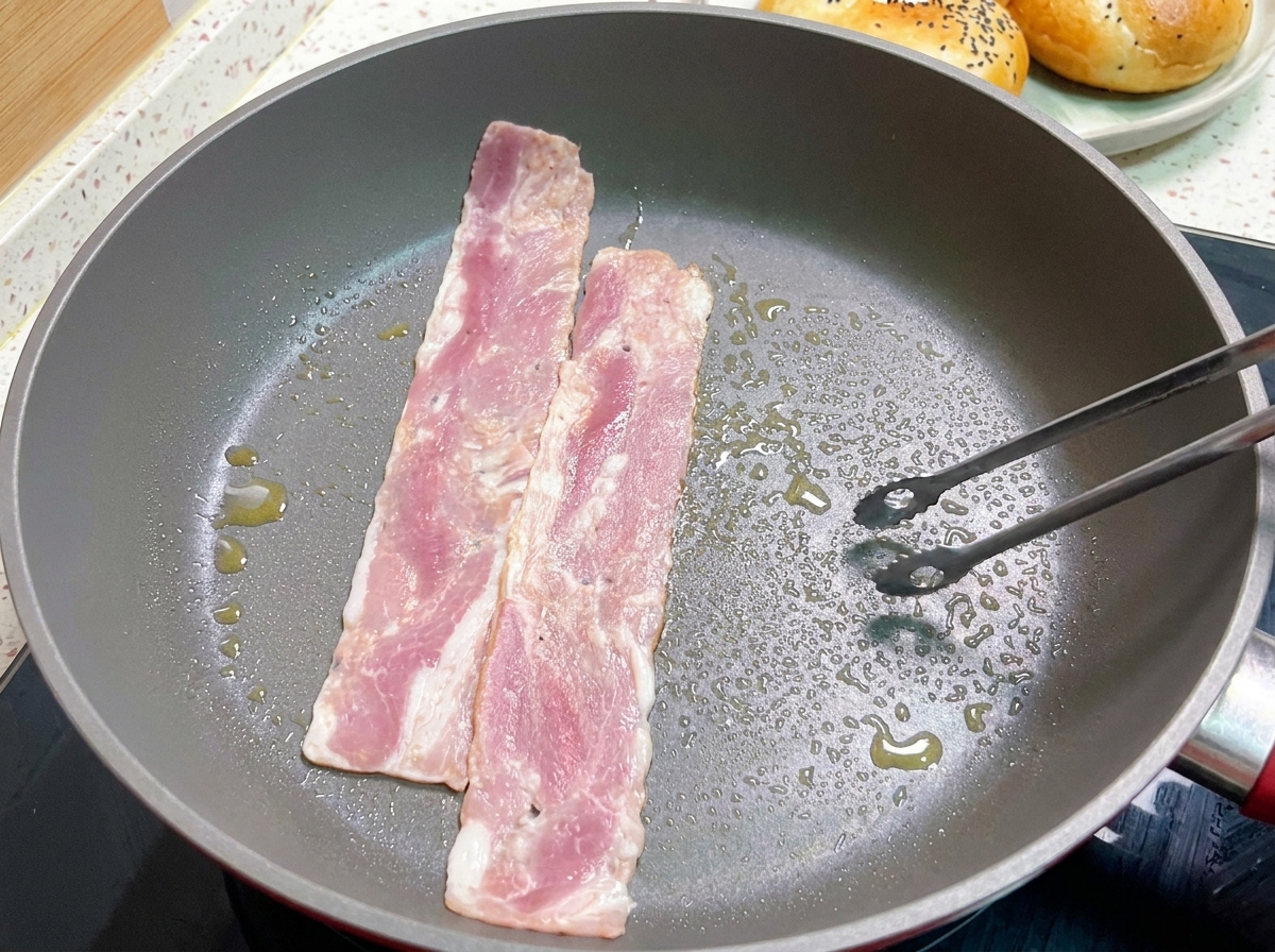 Strips of bacon frying in a hot pan, with tongs turning one of the lightly browned slices.