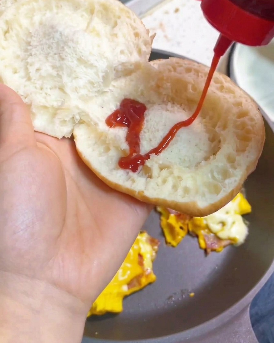 A hand squeezing a continuous zigzag pattern of bright red tomato ketchup from a bottle onto the inside of a toasted burger bun.