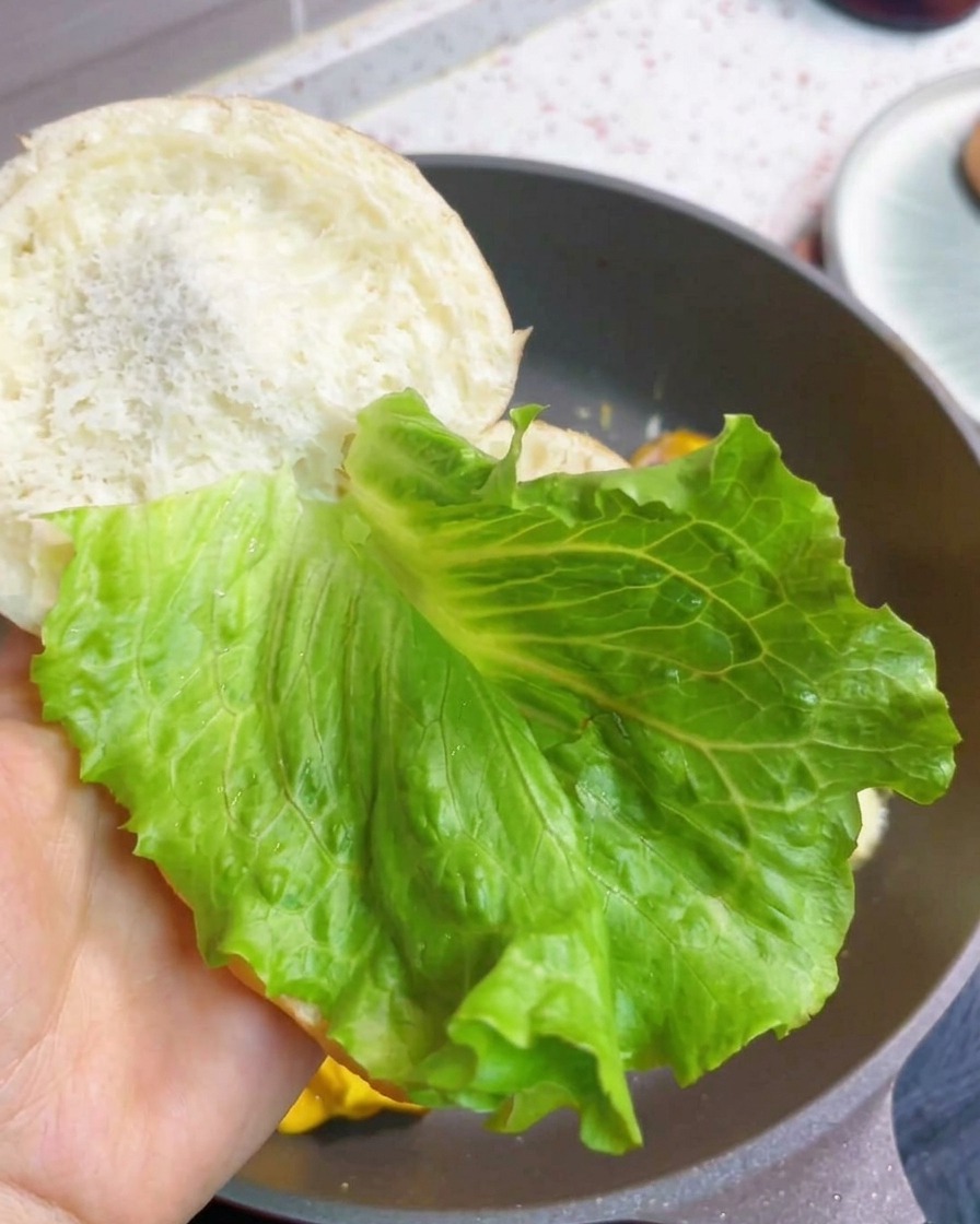 A breakfast burger being assembled with fresh lettuce and a cooked bacon-egg-cheese patty inside toasted buns.