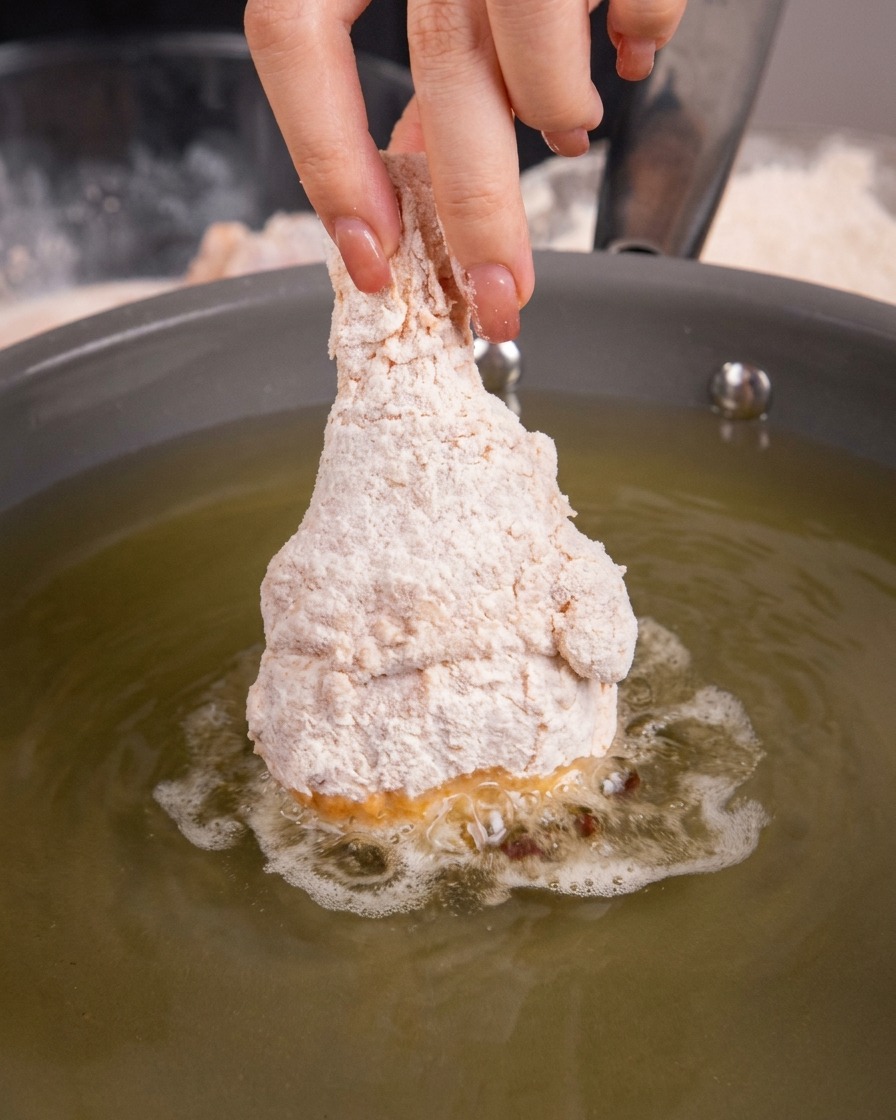 Breaded raw chicken drumstick being submerged into hot oil in a frying pan.