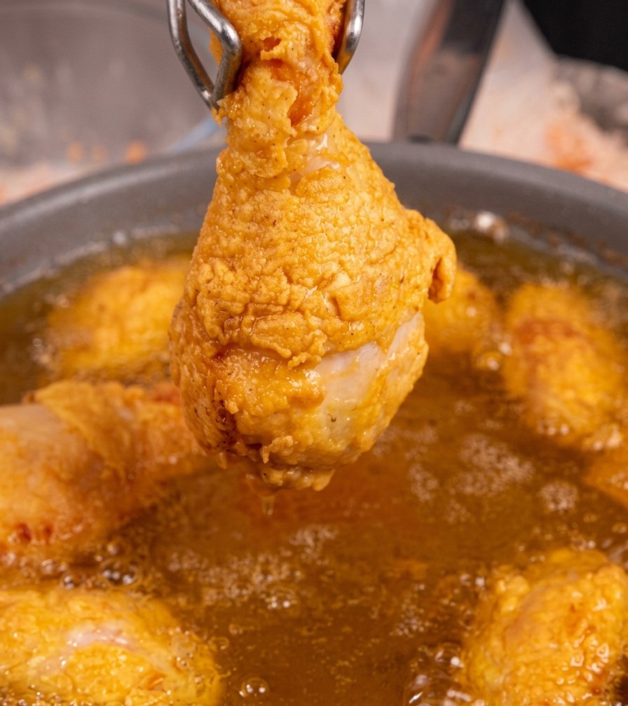 A single golden-brown fried chicken drumstick held by metal tongs over a pan of bubbling hot oil.