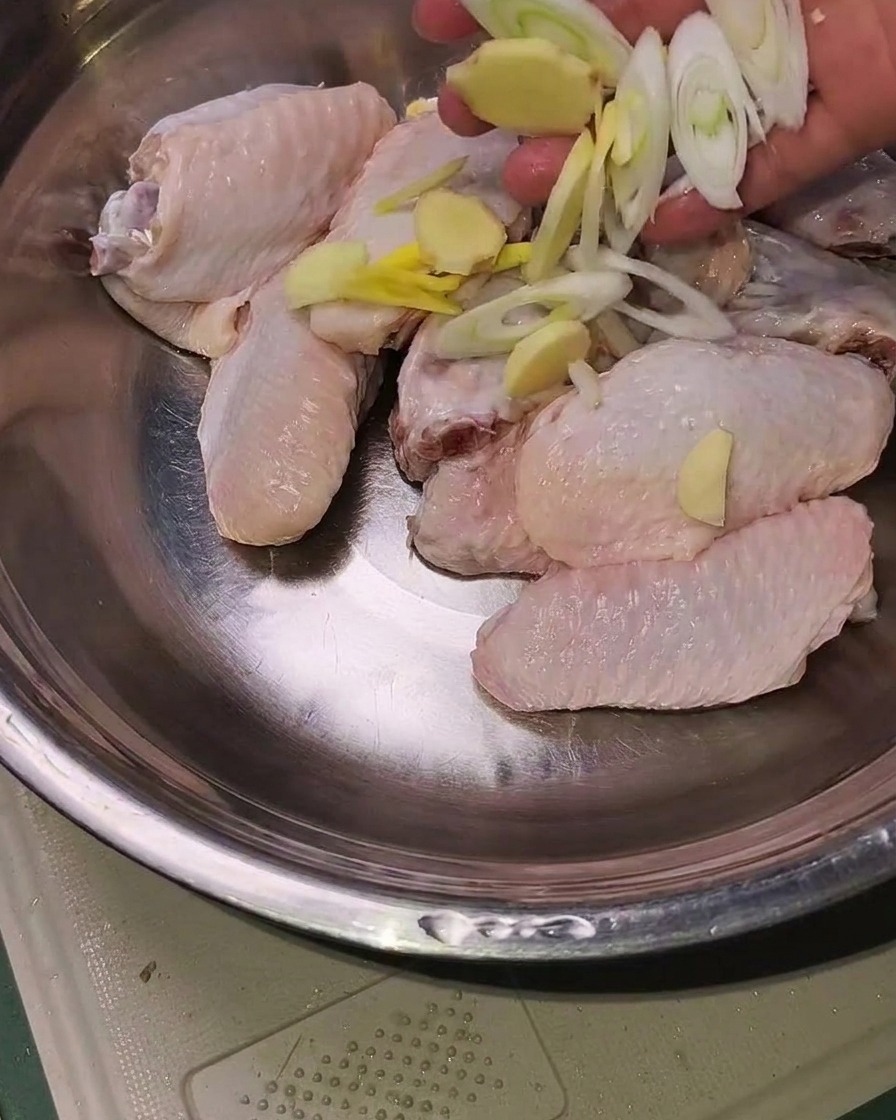 Raw chicken wings in a metal bowl topped with sliced ginger, garlic, and scallions ready for seasoning.