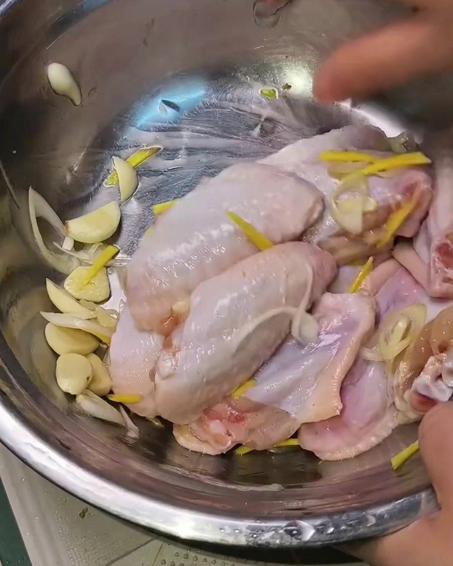Hands mixing the marinated chicken wings with aromatics in a metal bowl.