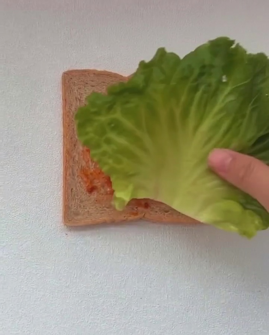 Two large green lettuce leaves being placed on top of a slice of toast already spread with red sauce.