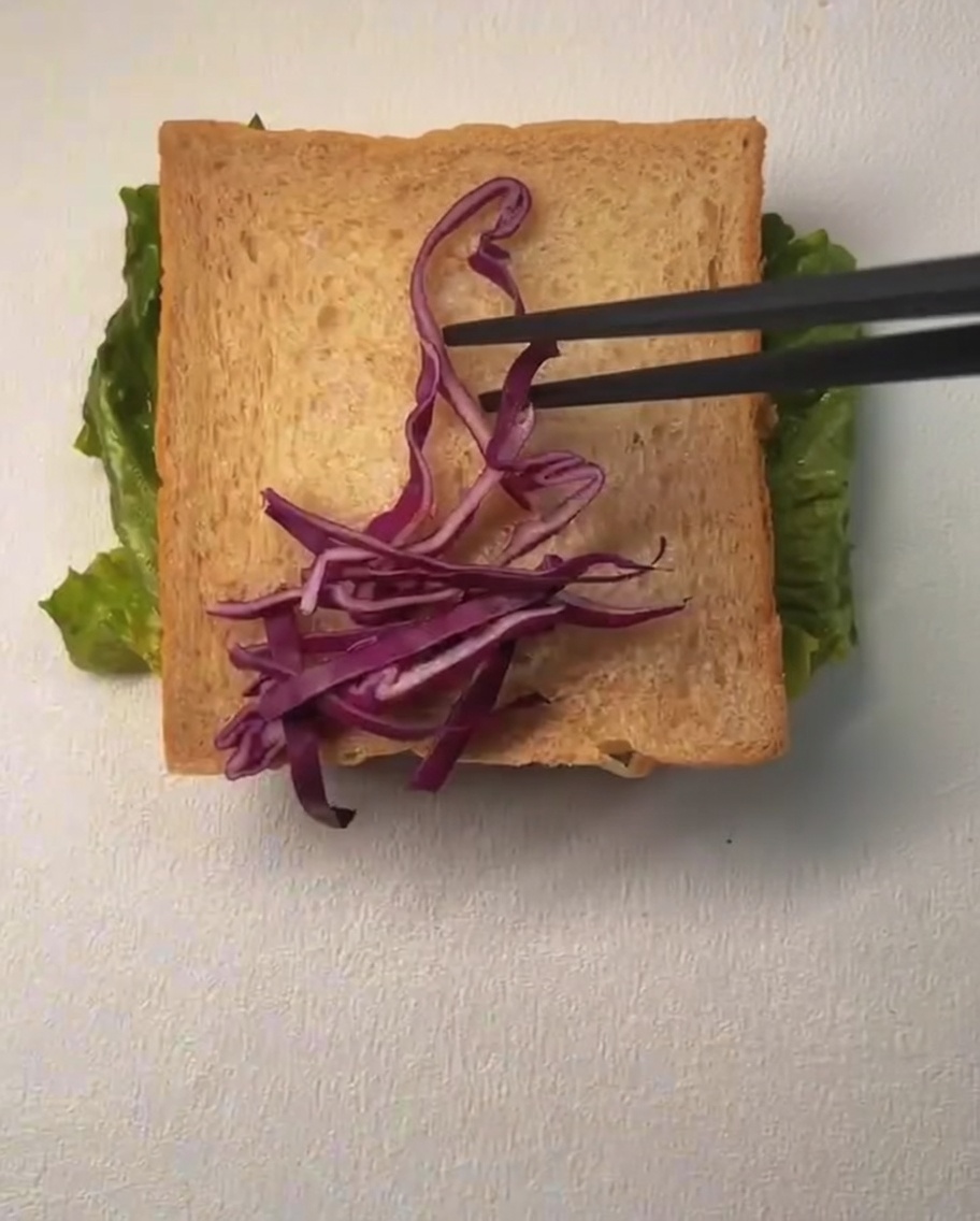 A slice of bread being topped with shredded purple cabbage and corn kernels, with mayonnaise being drizzled over the top.