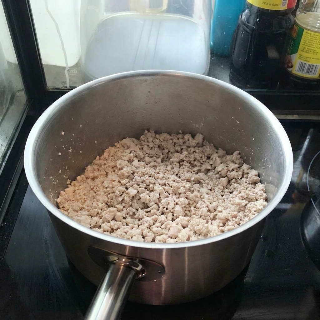 Minced pork browning in a stainless steel pot, stirred with a spatula.