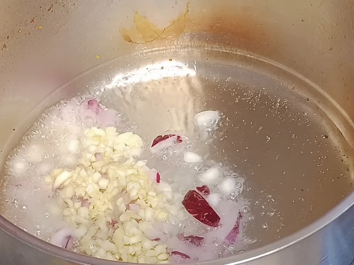 Chopped red onions and minced garlic simmering in hot oil inside a metal pot.