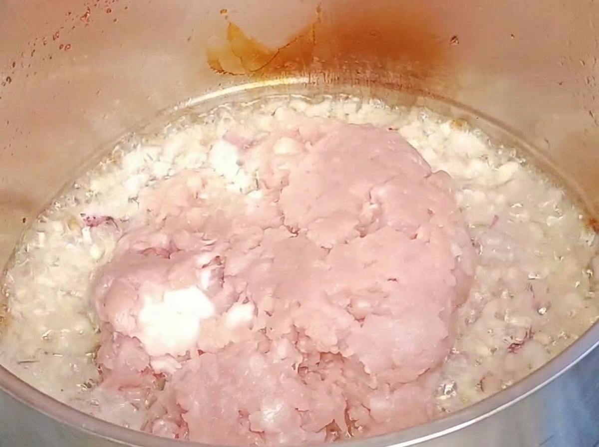 Raw ground meat being added over sautéed onions and garlic in a metal pot.