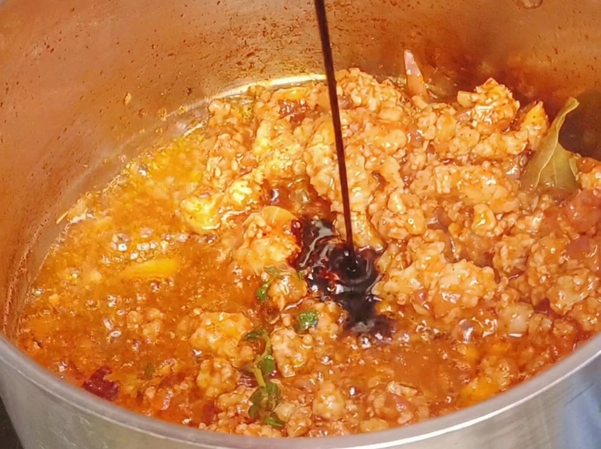 Dark soy sauce being poured into a pot of cooked, reddish ground meat.