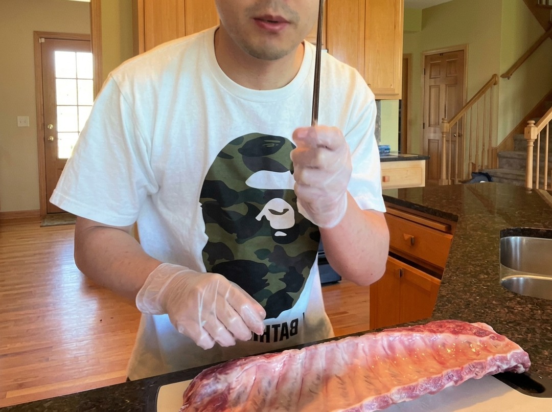 A close-up view of a chopstick being used to lift the membrane from the back of a rack of raw pork ribs.