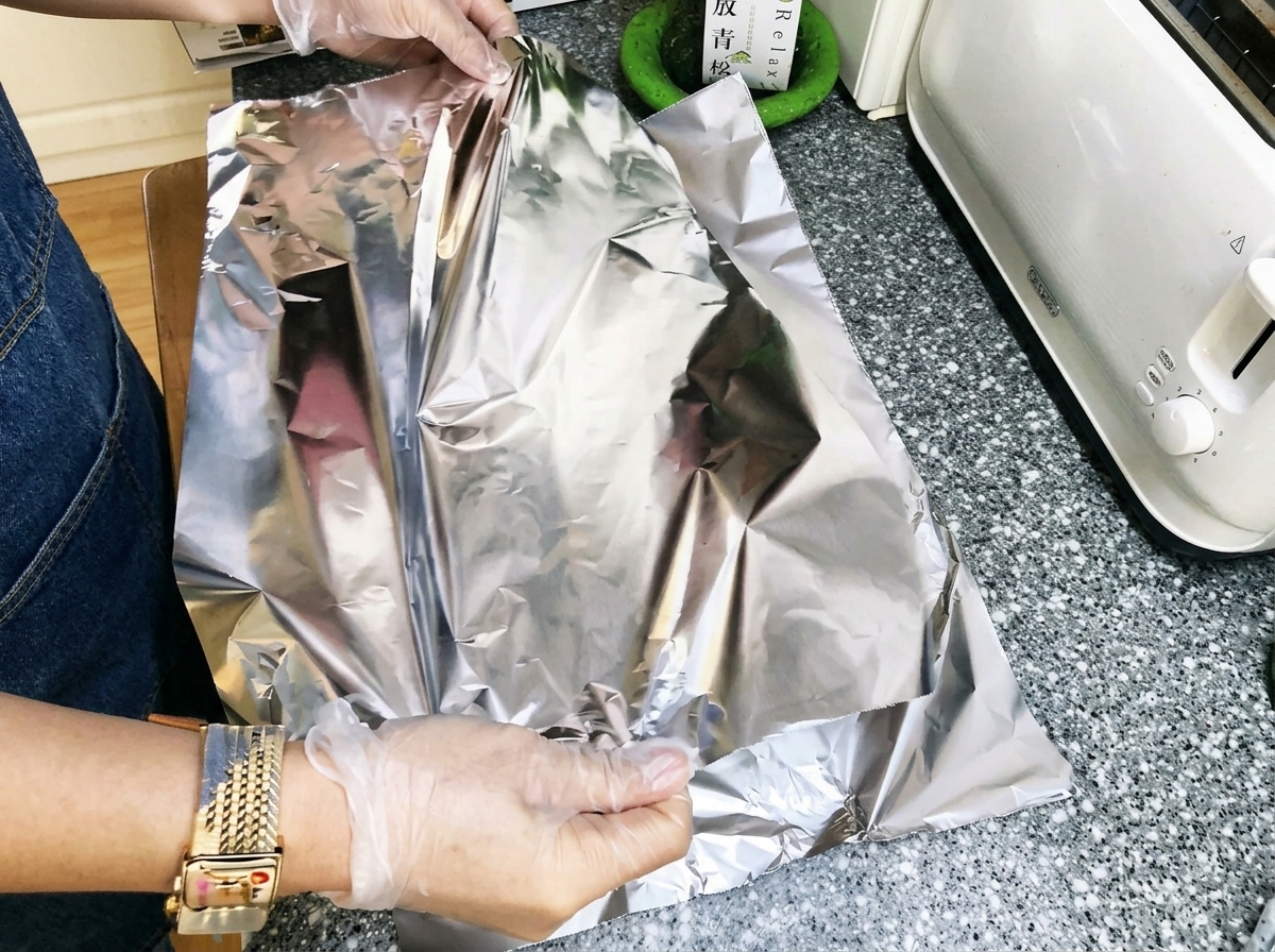 Hands folding aluminum foil tightly over marinated pork ribs to create a sealed package.