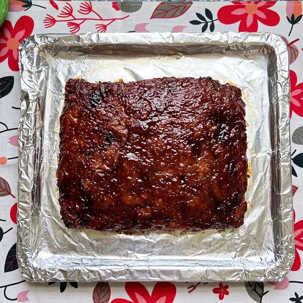 A close-up of a slab of cooked pork ribs on foil being coated with a thick, dark glossy sauce.