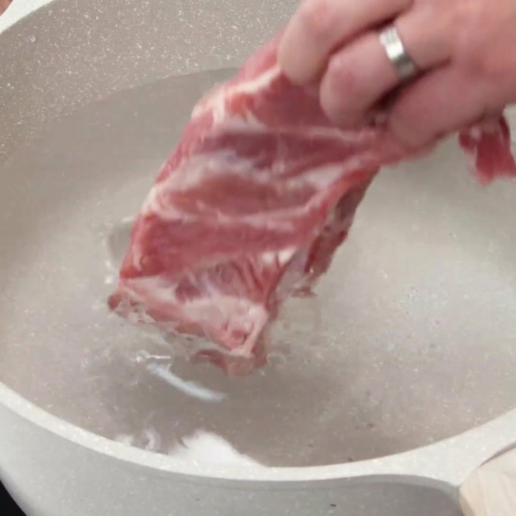 A hand placing a rack of raw pork ribs into a pot filled with cold water.