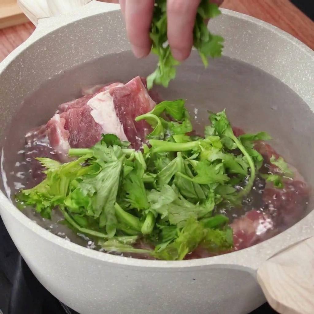 A hand dropping chopped celery leaves and aromatics into a pot of water containing raw pork ribs.