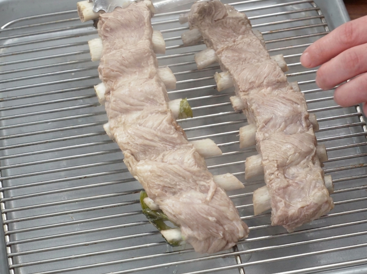 Boiled pork rib racks resting on a metal wire rack with a plate placed on top to flatten them.