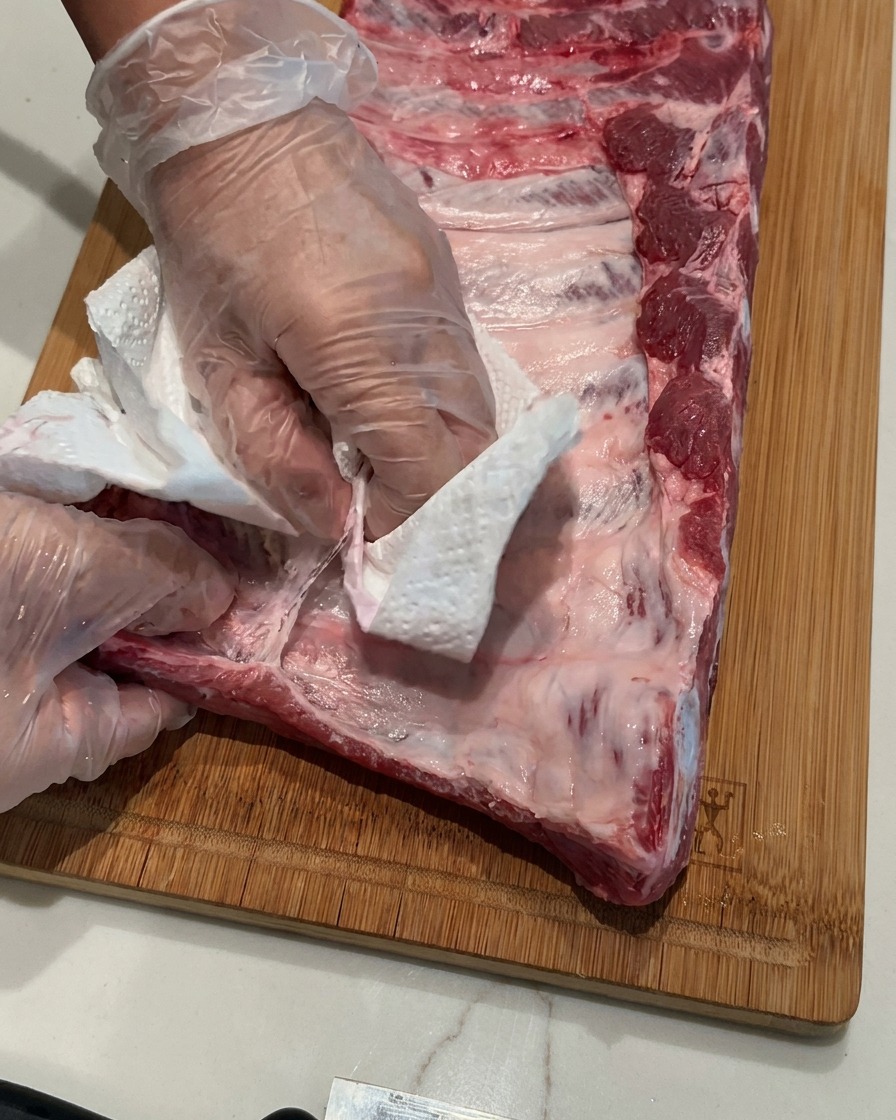 Hands wearing clear plastic gloves using a white paper towel to peel a thin membrane off a raw rack of pork ribs on a wooden cutting board.