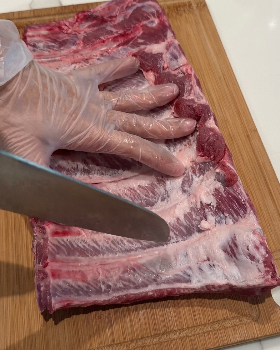 A gloved hand holding a chefs knife, using the tip to make small punctures in a raw rack of pork ribs.