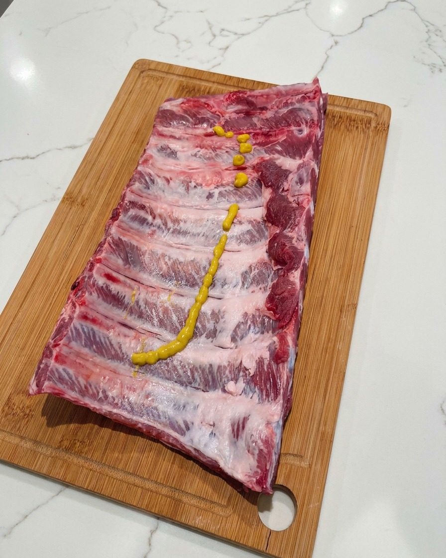 A squiggly line of yellow mustard squeezed over the top of a raw rack of pork ribs sitting on a wooden cutting board.