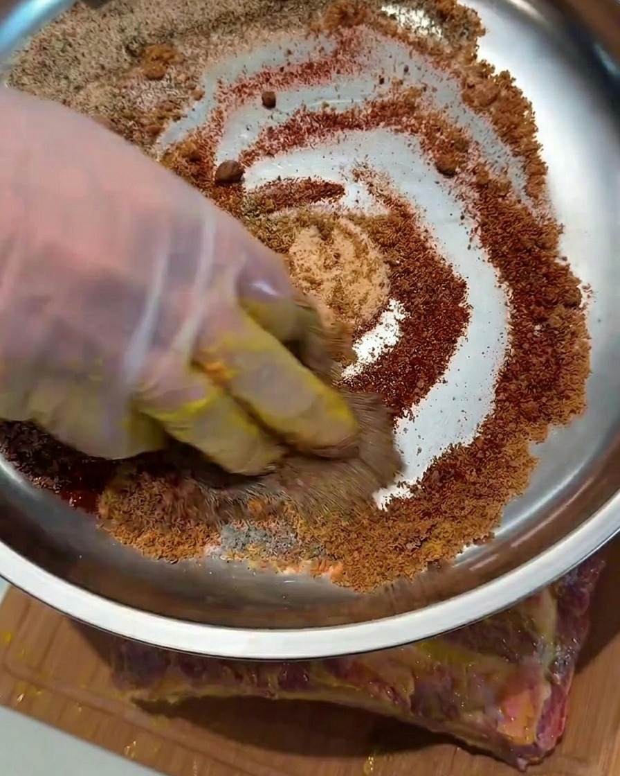 A close-up view of gloved hands pressing dry spices into the surface of mustard-coated pork ribs.