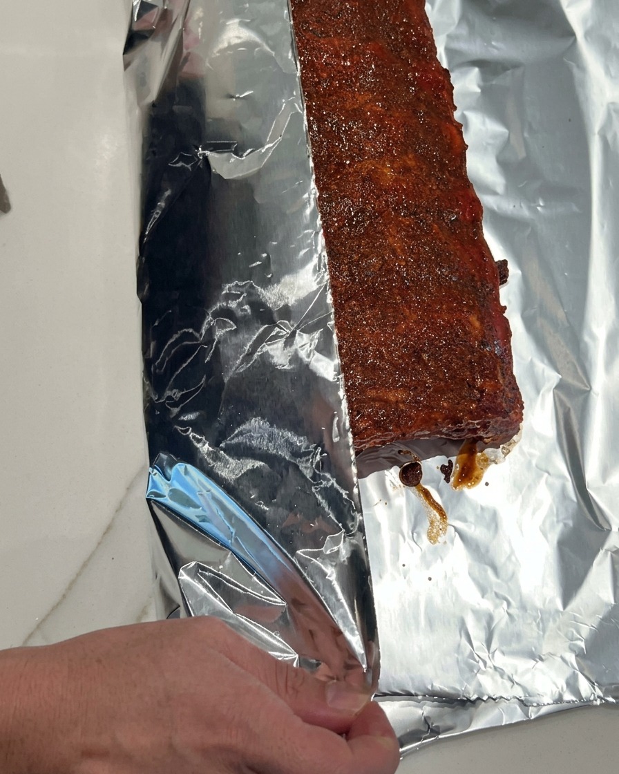 A hand carefully folding a large, shiny piece of aluminum foil over a sauced rack of pork ribs to seal them tightly.