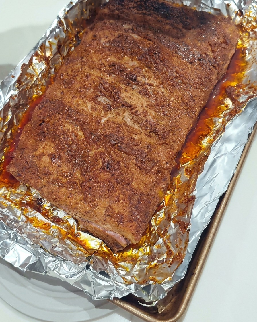 A silicone brush applying a glossy layer of thick BBQ sauce onto the surface of roasted pork ribs in a foil-lined pan.