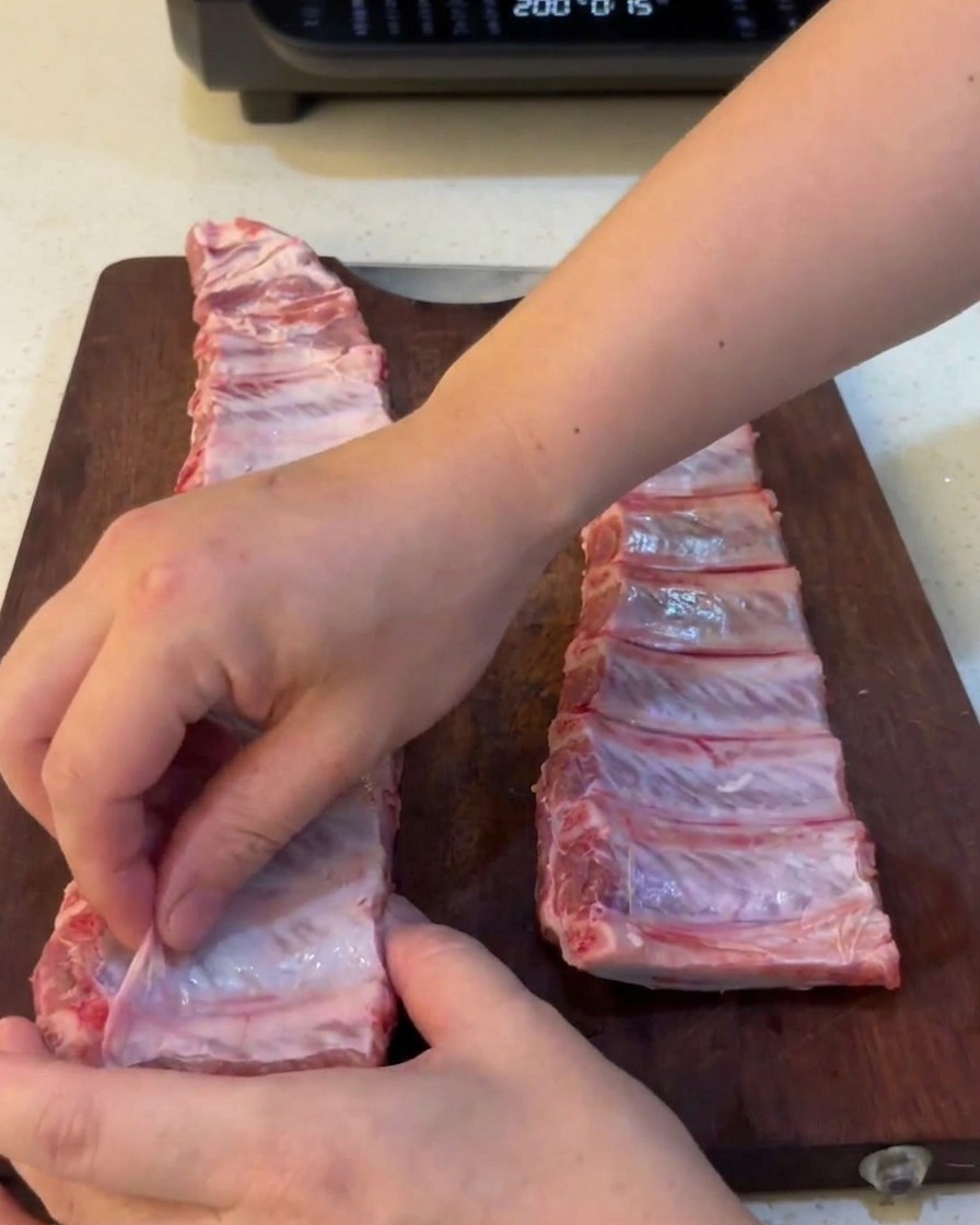 Close-up of hands pulling a thin, translucent membrane away from the back of raw pork ribs.
