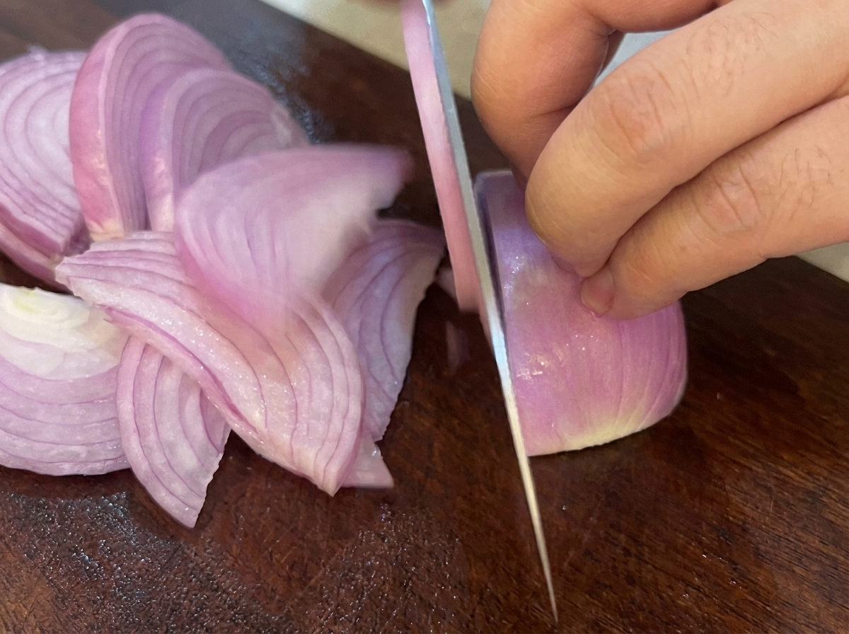 A knife slicing a red onion into strips on a dark wooden cutting board.