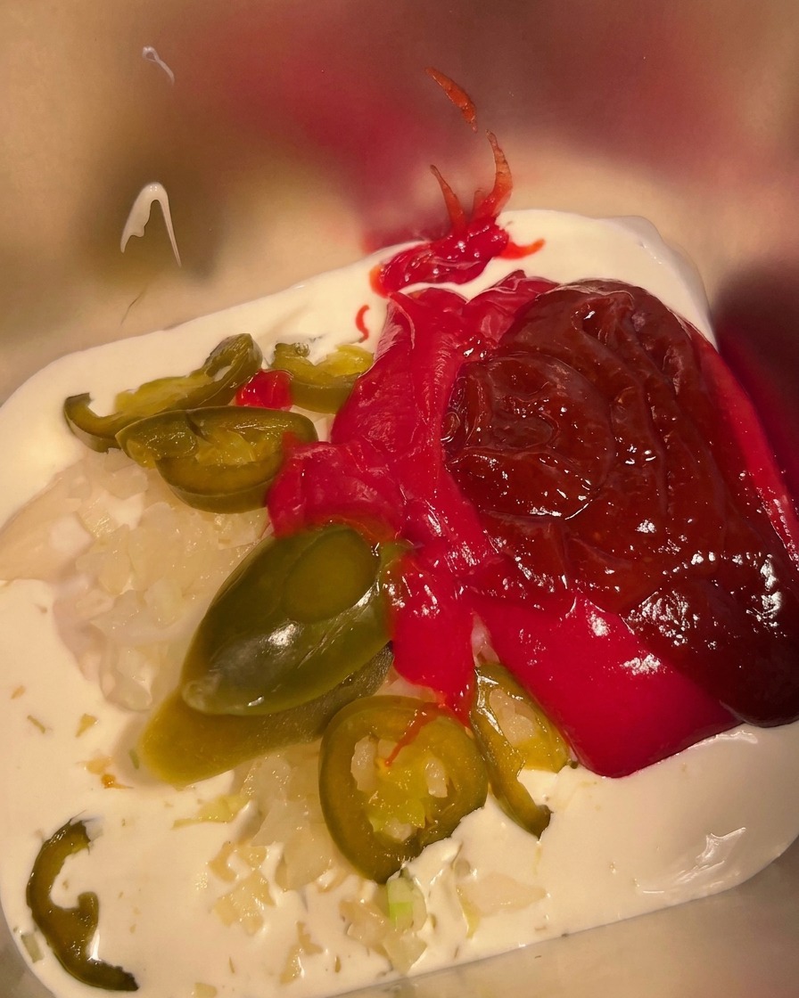 Chili powder being sprinkled over the mixture of mayonnaise, cream, and hot sauce in a metal container.