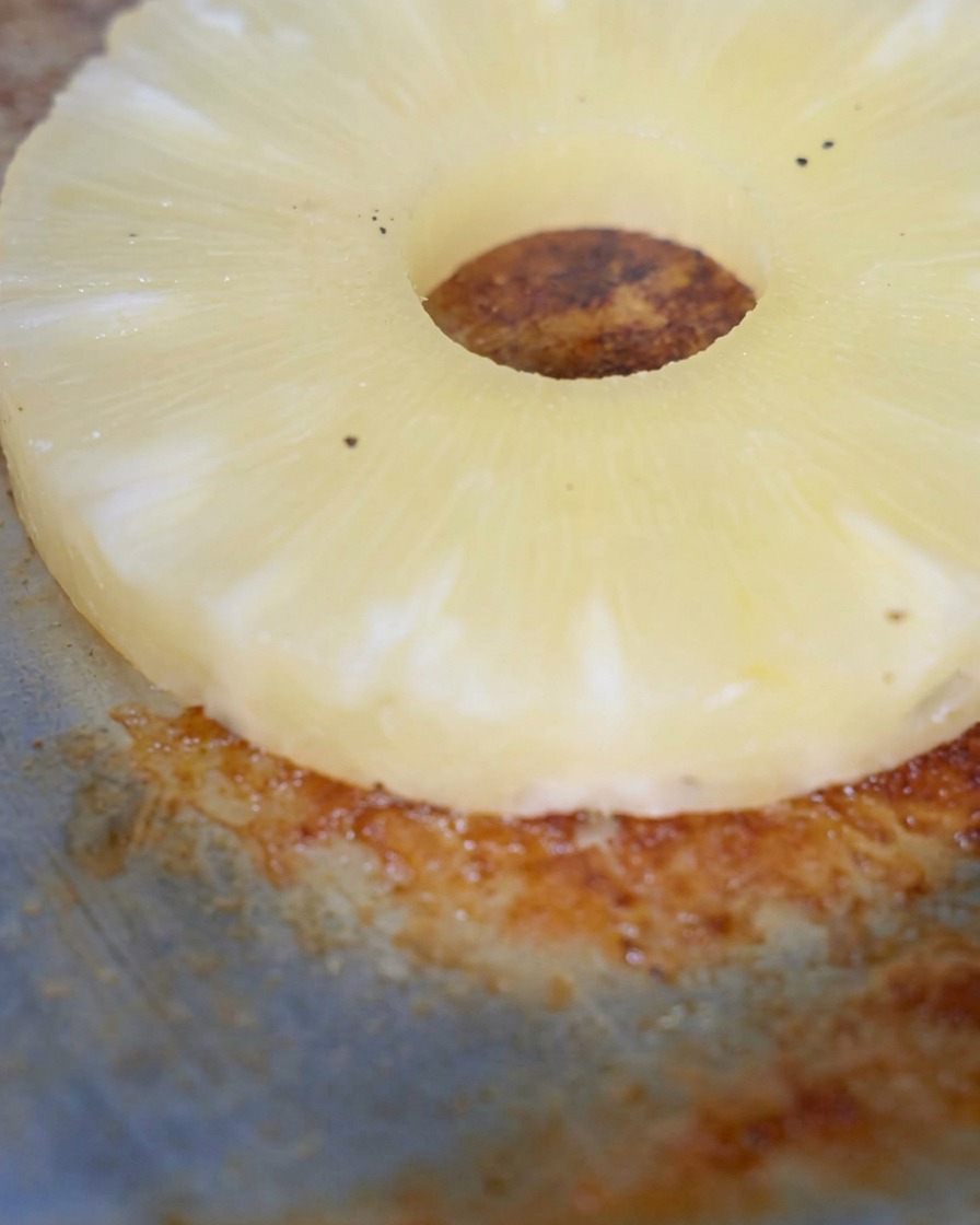A single slice of pineapple grilling on a hot, dark griddle surface.