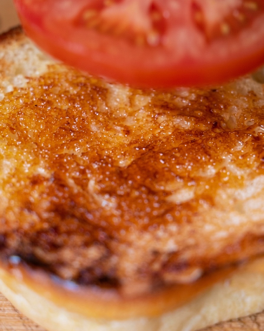 A slice of fresh tomato being placed on the bottom toasted burger bun.