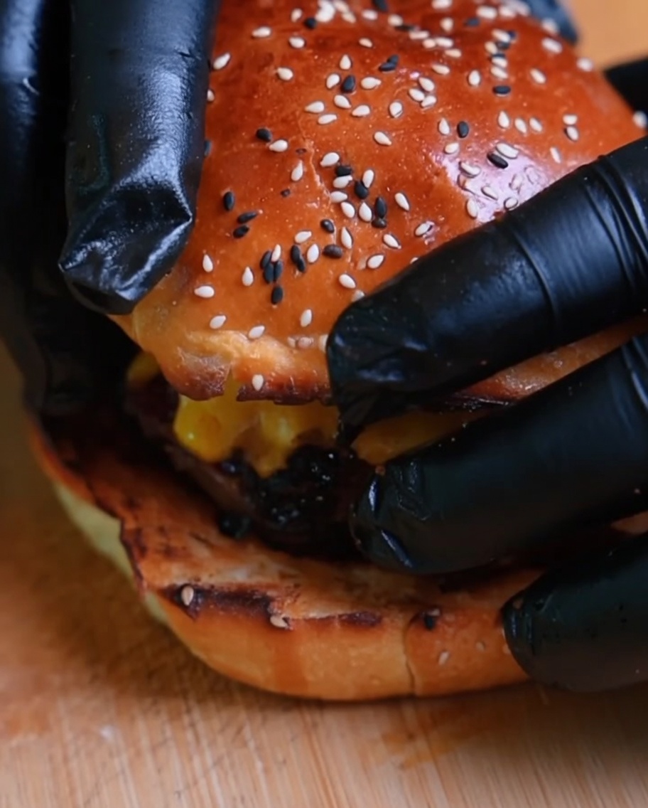 A close-up of a burger being finished with the top bun placed on it, held by gloved hands.