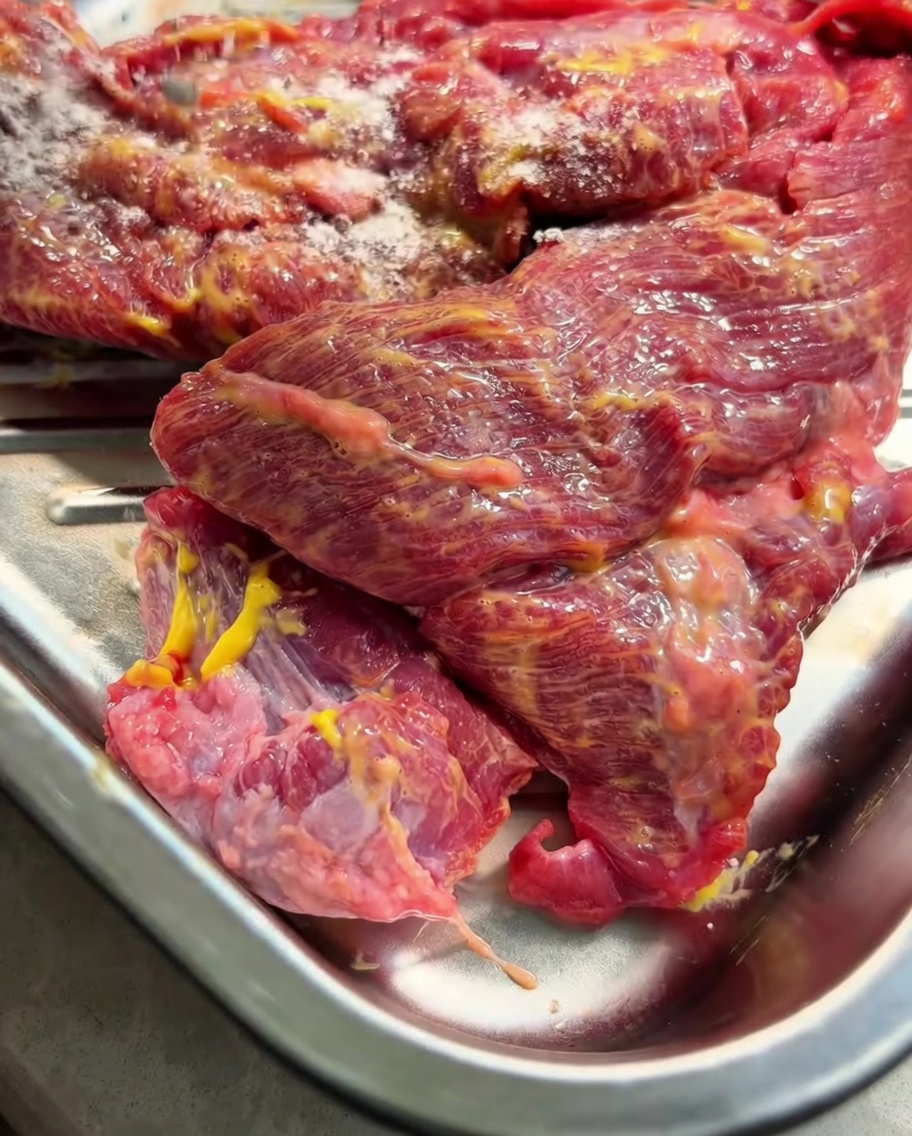 The mustard-coated beef brisket in a metal tray, with dry seasoning powder being sprinkled over the top.
