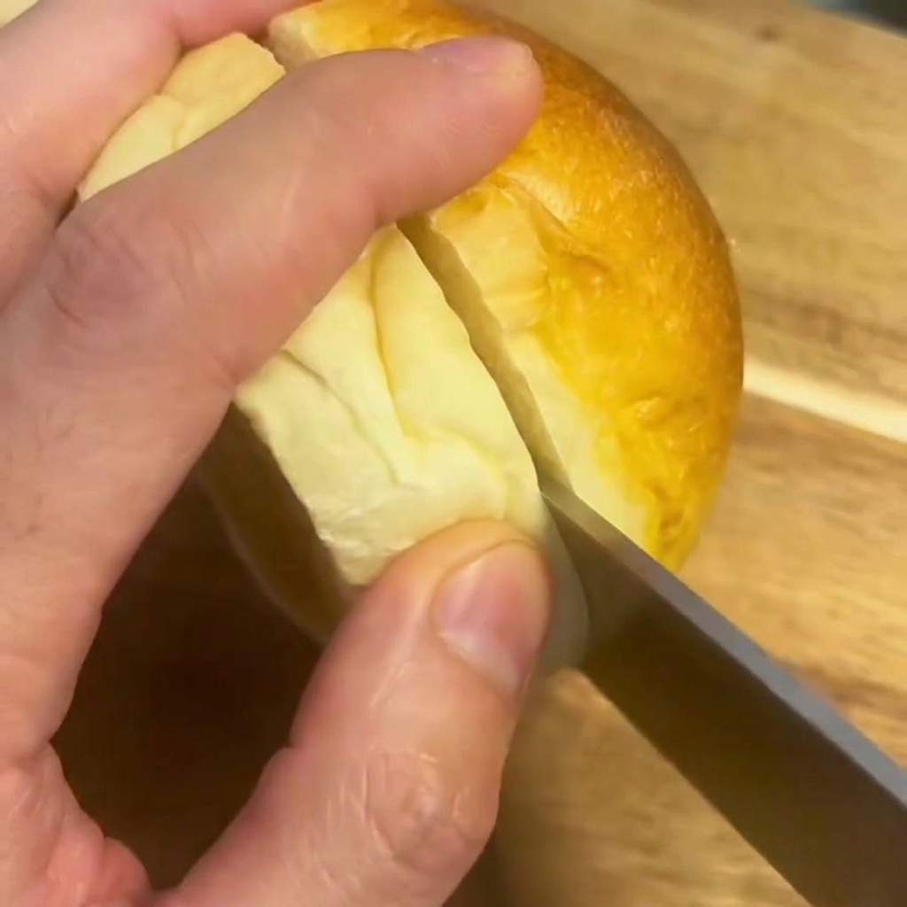 A knife cutting a soft burger bun in half on a wooden cutting board.