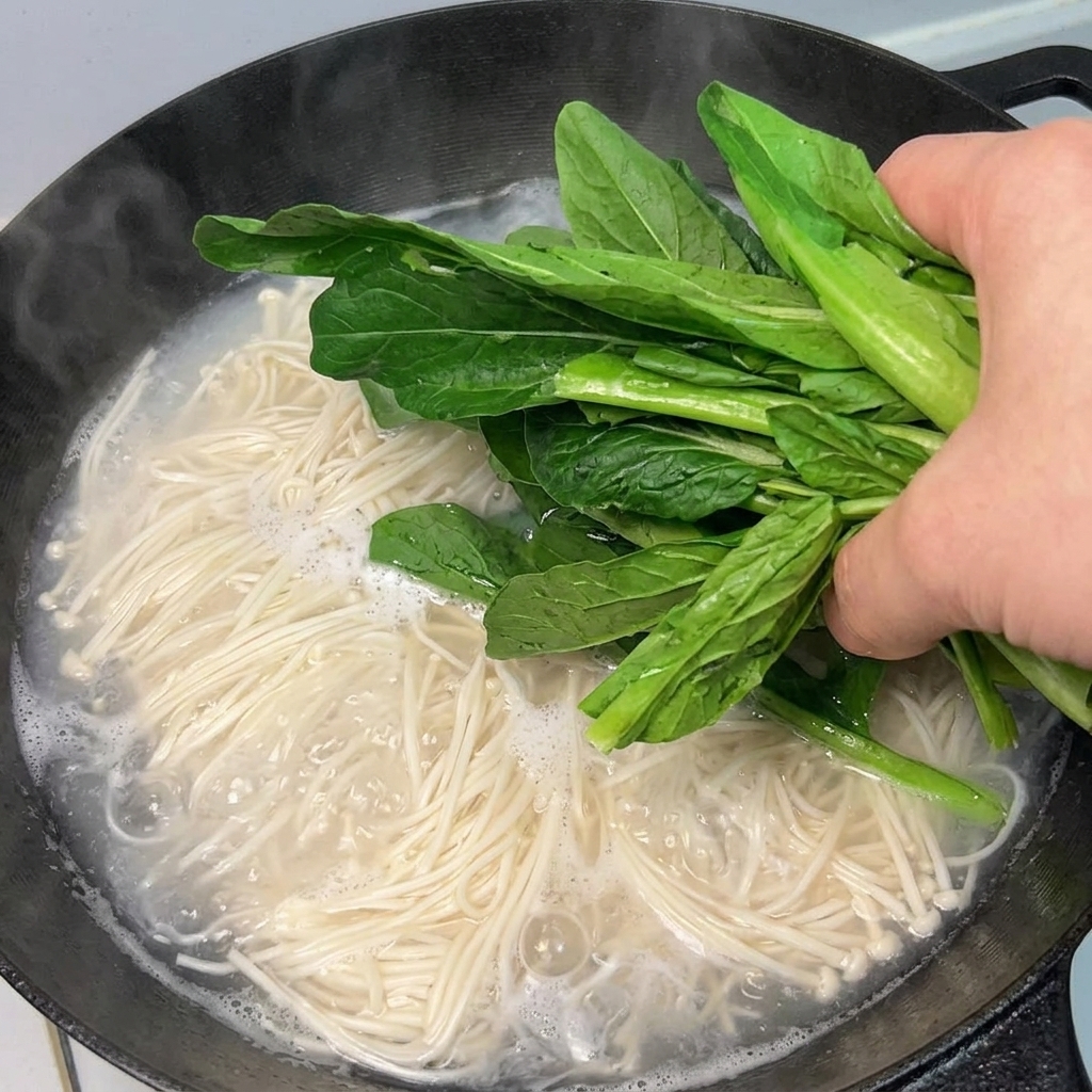 Fresh green leafy vegetables being added to a pot of boiling noodles.