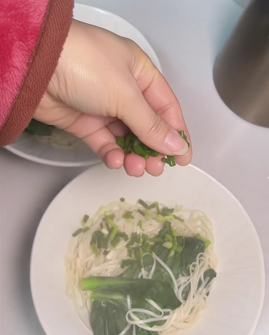 Hand sprinkling chopped green onions over a bowl of noodles.