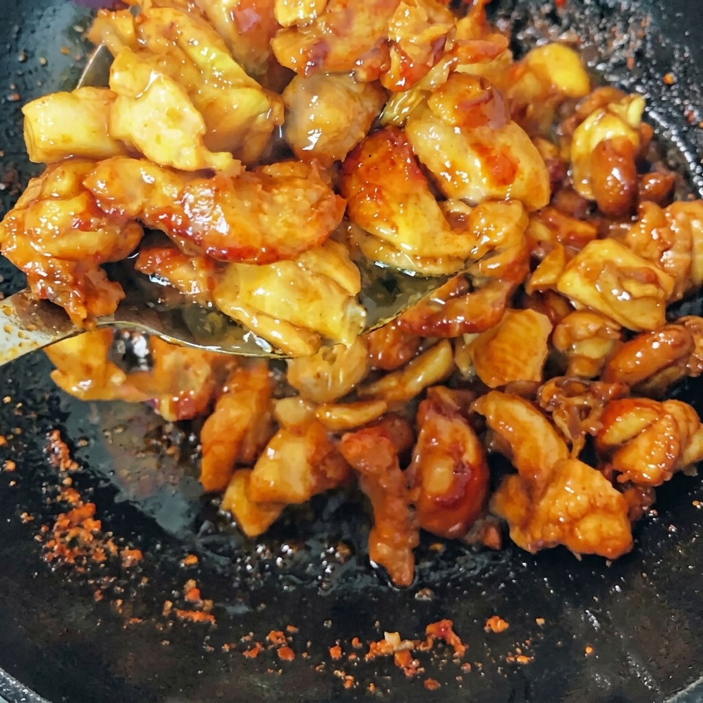 Browned chicken pieces stir-frying in a wok with green onions, ginger slices, and dried chili peppers.