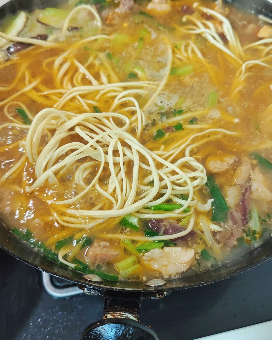 Green vegetables and chopped garlic sprouts being stirred into the boiling noodle soup.