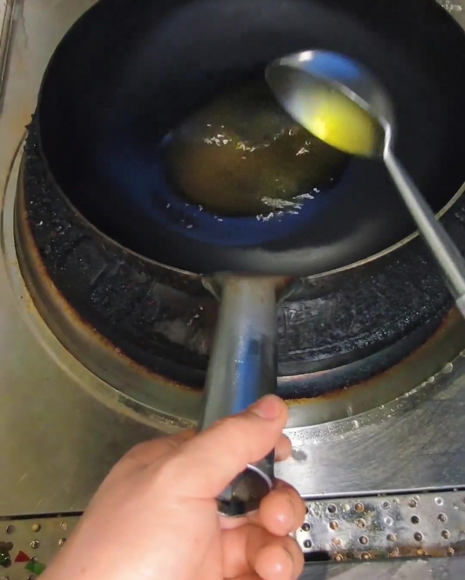 A metal ladle pouring yellow cooking oil into a dark hot wok.
