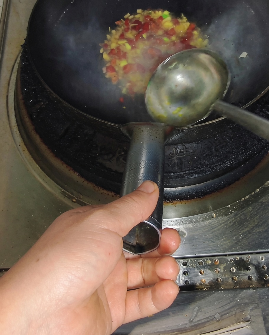 A ladle pouring clear broth into a hot wok containing sautéed diced chilies and green onions, producing steam.