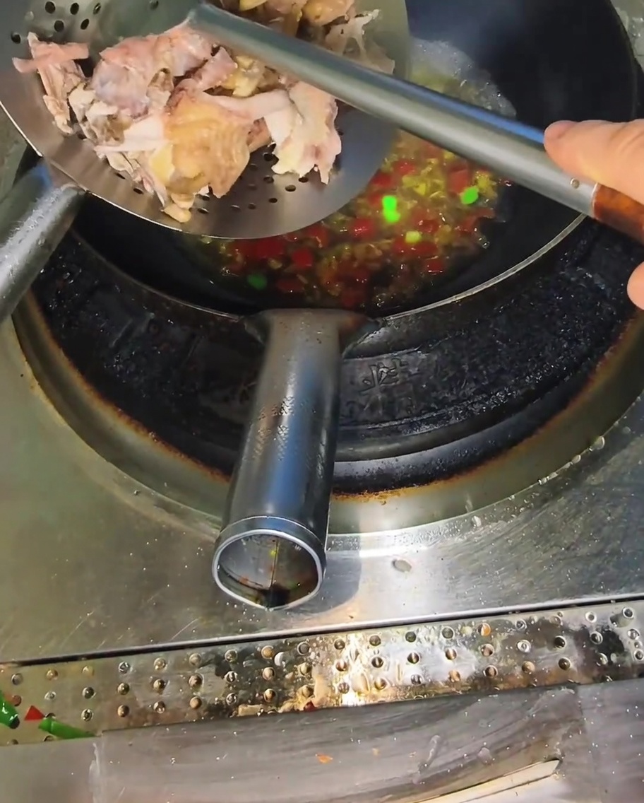 A metal slotted spoon adding pieces of cooked, shredded chicken into a wok filled with a bubbling spicy broth.