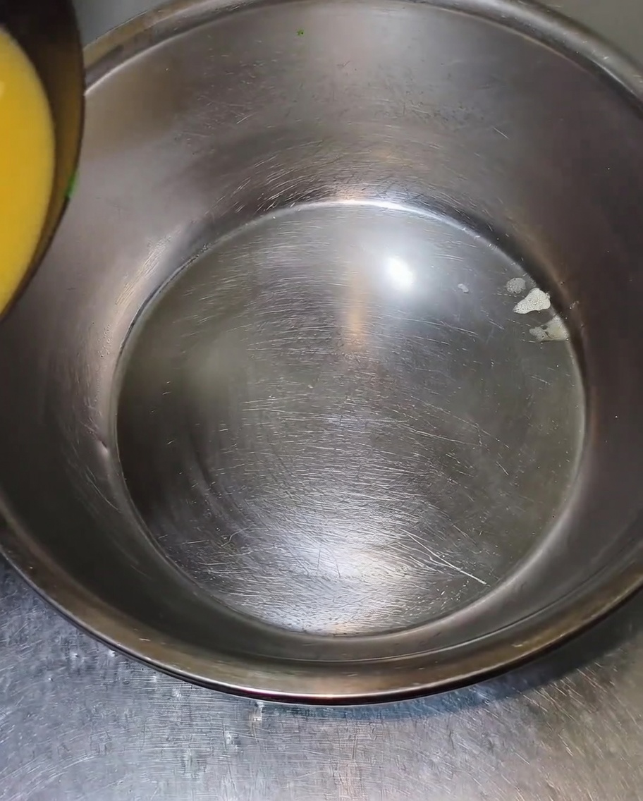 Hot, yellowish broth being poured from the edge into a large, empty stainless steel mixing bowl.