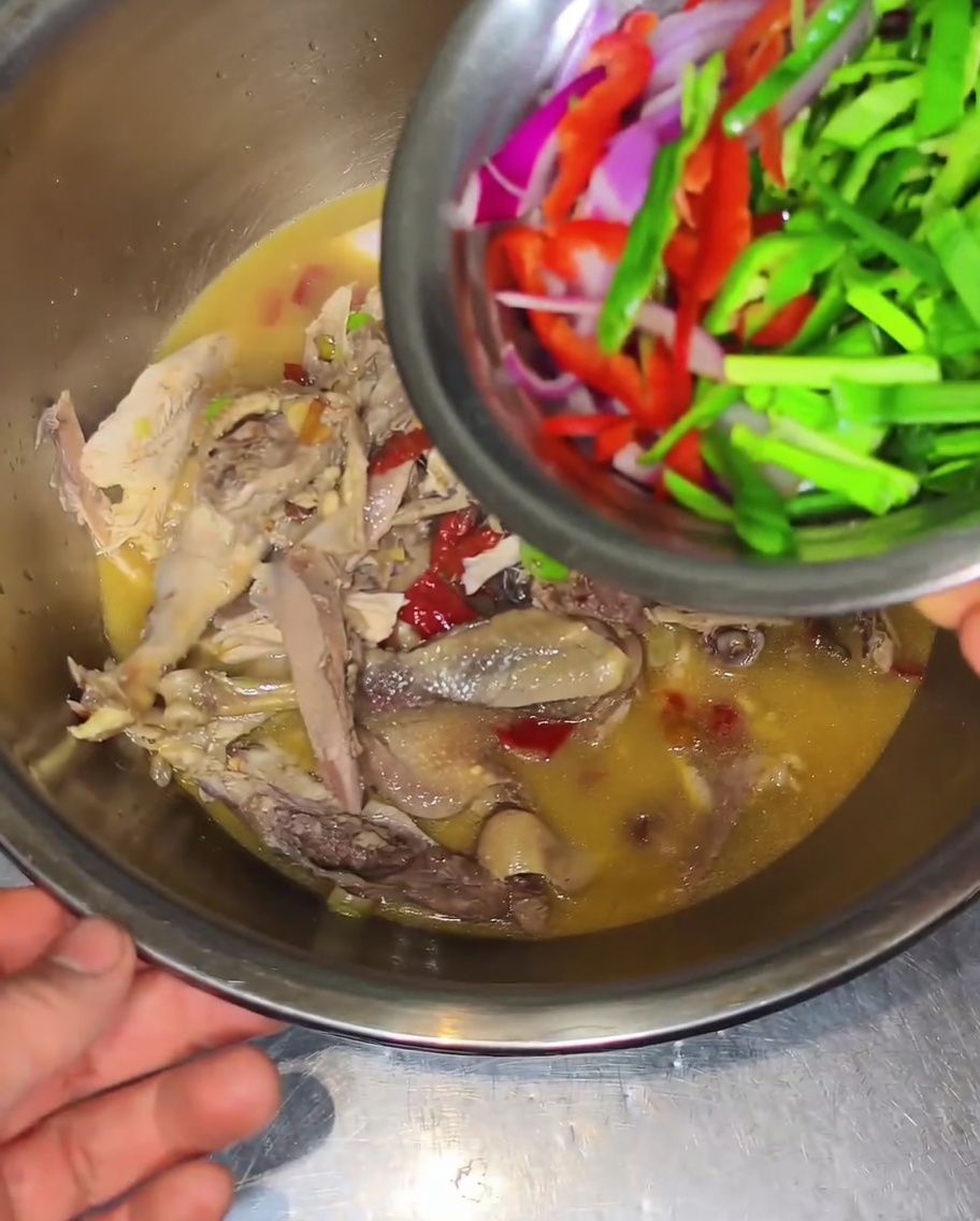 A small metal bowl pouring thinly sliced red onions, green peppers, and red peppers over cooked chicken in broth in a large mixing bowl.